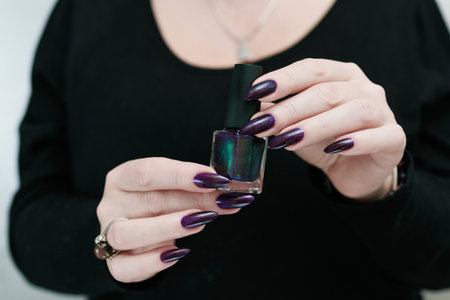 Female hands with long nails and black purple manicure holding a bottle of nail polishの写真素材