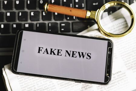 Smartphone with the phrase fake news written on the screen on a laptop, newspaper and magnifying glass Conceptual of hoaxes and fake news through new technologiesの写真素材