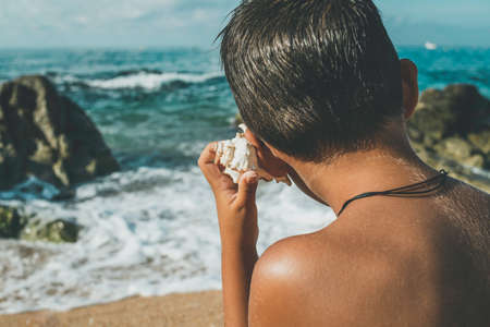 Child listens to the sound of the sea with a conch shell in front of the beachの写真素材
