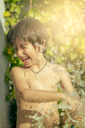 Laughing boy in swimsuit plays and gets wet with water from a hose at sunsetの写真素材