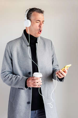 Stylish mature man in gray jacket listens to hard rock on his smart phone while drinking coffee. Conceptual of lifestyleの写真素材