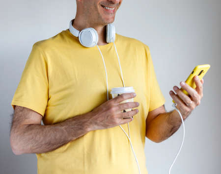 Man in yellow shirt and headphones drinks coffee while checking his smart phoneの写真素材
