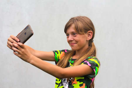 A ten year old girl poses and takes a selfie with her hand on her phone in a brown case. Gray white background. Smiling attractive girl is having fun at the camera in colorful clothes Selective focus.の写真素材