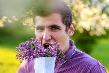 Defocus close-up portrait of handsome caucasian man with cone flowers in lilac garden. Funny attractive brunette guy smelling fresh flowers and looking at camera on blurred background. Out of focus.の写真素材