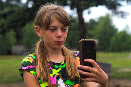 Defocus surprised ten year old girl find something on smart phone. Beautiful teenage girl takes selfie while standing alone on park. Blogging and live streaming. Generation z. Out of focus.の写真素材
