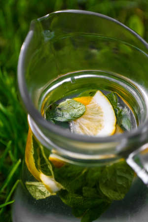 Defocus close-up slice lemon and leaves of mint in glass jug of lemonade natural green background. Pitcher of cool summer cocktail or punch outdoor, outside. Sunny summer day. Heat. Out of focusの写真素材
