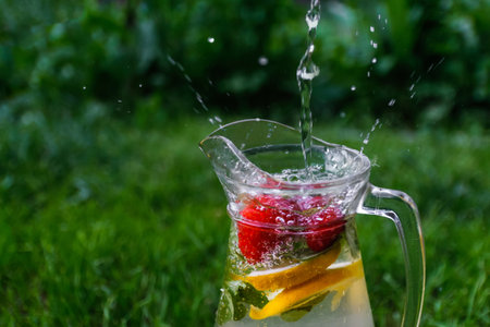 Defocus jet splash spray water bubbles in glass jug of lemonade with lemon, strawberry and mint on natural green background with water drops. Pitcher of summer citrus drink. Copy space. Out of focusの写真素材