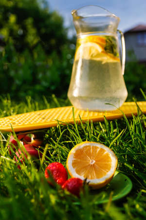 Defocus saucer with lemon and two strawberry standing on grass near jug of lemonade with slice lemon and leaves of mint on yellow board. Blurred green background. Fresh summer. Nobody. Out of focusの写真素材
