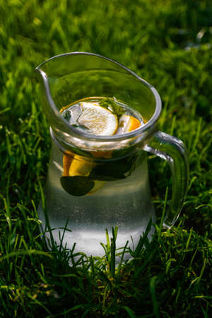 Defocus close-up glass jug of lemonade with lemon and mint on natural green nature background. Pitcher of cold summer cocktail or punch outdoor, outside. Drink mojito. Vertical. Out of focus.の写真素材