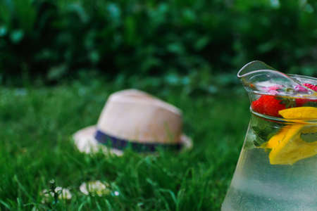 Defocus glass jug of lemonade with strawberry, slice lemon and leaves of mint in on blurred grass background. Pitcher of cool fresh cold drink. Side view. Hat and sunglasses. Out of focus.の写真素材