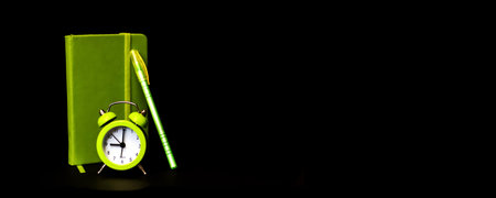 Light green alarm clock with pen notebook on black dark background. Top view. Wake up alert concept. Morning routine. Back to school. Minimalism. Selective focus.の写真素材