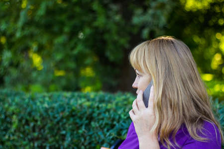 Defocus wonder caucasian blond woman talking, speaking on the phone outside, outdoor. 40s years old woman in purple blouse in park. Adult person using phone. Vertical. Out of focus.の写真素材