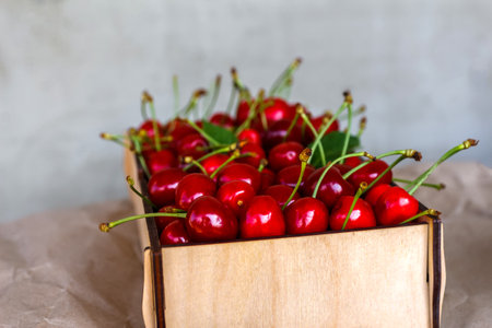 Cropped wooden box, crape of sweet red cherries with tail standing on craft yellow wrinkled paper. Stone concrete gray background. Summer fruits and berries. Harvest and crop concept. Organic foodの写真素材