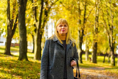 Portrait of gorgeous middle aged woman in autumn park. Beautiful woman in gray sweater and leather boots sitting under tree in autumn park with fallen leaves. Selective focus.の写真素材