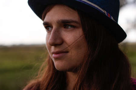 Defocus overall portrait of a serious young woman with brown hair wearing a hat outdoors. Green nature trees background, forest. Looking at camera. Out of focus.の写真素材