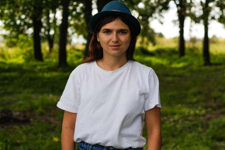 Defocus overall portrait of a serious young woman with brown hair wearing a hat outdoors. Green...