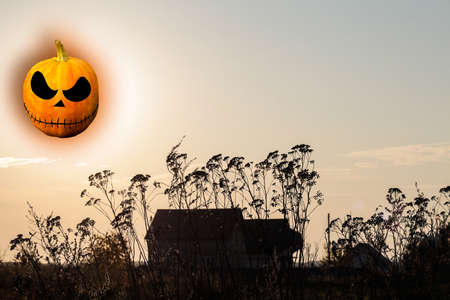 Halloween concept. Mysterious abandoned house at sunset. transparent Pumpkin Jack lantern instead of the moon and sun. Weeds are in the foreground. Terror and fear concept. Selective focus.の写真素材