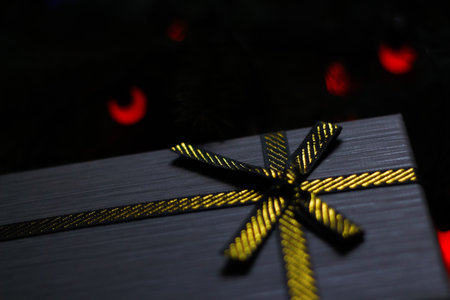 Close-up christmas gray present box with golden ribbon fir-tree branches on the dark background close-up. Side view. Red bokeh and light spot. Dark mood. Selective focus.の写真素材