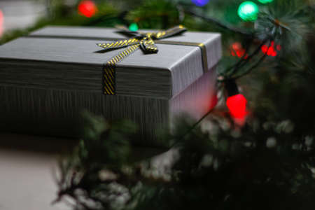 Defocus gray gift with gold bow ribbon on natural fir tree blurred background with glowing festive green, red light bulb at night. Christmas present. Holiday box. Out of focus.の写真素材