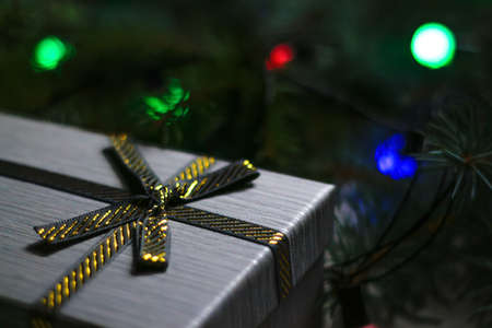 Defocus gray gift with close-up gold bow ribbon on pine or fir tree blurred background with glowing festive green, red and blue light bulb at night. Christmas present. Holiday box. Out of focus.の写真素材