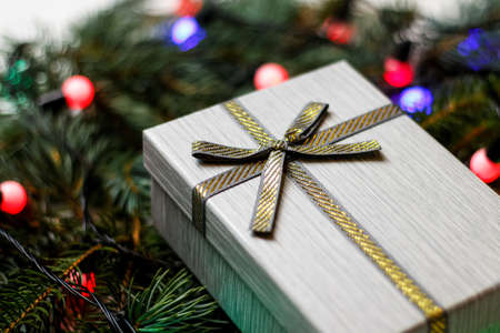 Defocus gray gift with gold bow on pine ir fir tree blurred background with glowing festive light bulb. Christmas present background. Holiday box. Out of focus.の写真素材