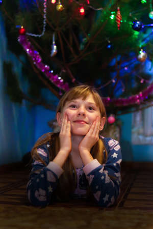Defocus little blonde Caucasian girl 10 years old in pajamas lies near the Christmas tree She put her head in her hands. Dark shadow The concept of sleep, daydreaming, dreaming, desires. Out of focus.の写真素材