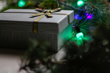 Defocus gray gift with gold bow ribbon on natural fir tree blurred background with glowing festive...