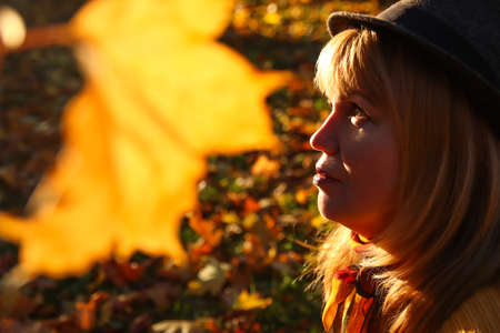 Close-up portrait of pretty Caucasian blonde woman in colorful orange autumn park. Bright stylish woman in hat and knitted pullover near tree trunk. Hello fall concept. Yellow maple leaf. Love nature.の写真素材