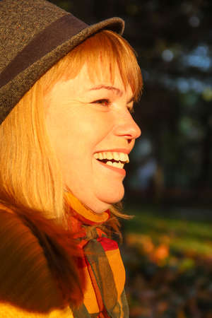 Close-up portrait of pretty Caucasian blonde woman in colorful orange autumn park. Bright stylish woman in hat and knitted pullover near tree trunk. Hello fall concept. Yellow maple leaf. Love nature.の写真素材