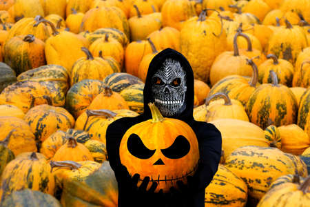 Grim reaper holding halloween pumpkin head. Man in death mask with fire flame in eyes on many orange pumpkins background. Halloween holiday concept. Dark horror.の写真素材