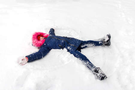Preteen happy girl in bright pink warm hood making snow angel while lying on snow outside background. Pretty child outdoor, cold weather. Caucasian kid. Teenage riot. Winter joy.の写真素材