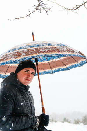 Close-up portrait of young man in warm hat outside on rural winter snowy house background. Happy millennial smiling outdoor, cold weather. Caucasian guy 20-25 years. Face with acne or herpes.の写真素材