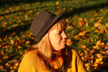 Portrait of young Caucasian blonde woman in autumn park. Bright stylish woman in yellow pullover and shawl of neck seating in october park. Happy fall concept. Sunny weather, sunset.の写真素材