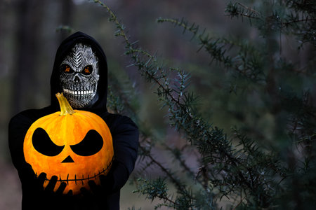 Grim reaper holding halloween pumpkin head. Man in death mask with fire flame in eyes on dark black nature juniper plant background. Halloween holiday concept. Dark horror.の写真素材