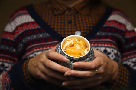 Closeup of female hands with a mug of creative beverage of mandarin orange lobule. Young hipster woman in Christmas sweater smelling cup of fruits. Cozy warm ceramic. Long nails. Winter detail.の写真素材