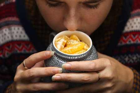 Closeup of female hands with a mug of creative beverage of mandarin orange lobule. Young hipster woman in Christmas sweater smelling cup of fruits. Cozy warm ceramic. Long nails. Winter detail.の写真素材