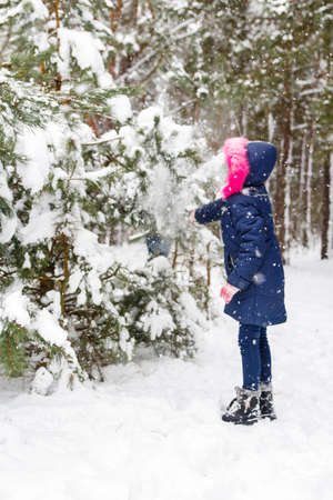 Preteen happy girl in pink warm hood shakes the snow from a branch outside on nature winter snowy forest background. Pretty child outdoor, cold weather. Caucasian kid. Walking on woodland. Closeup.の写真素材