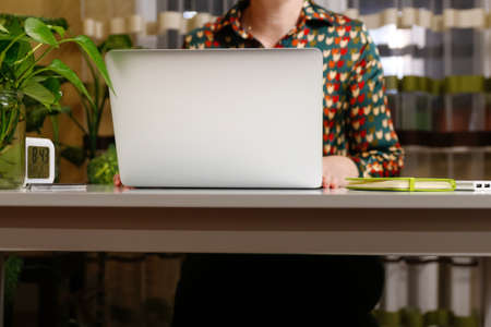 Unrecognizable woman with working on computer at home. Start or finish of a work day. Young woman seating at the working table and laptop. Workplace. Female hand. Green notebook, plant. Copy space.の写真素材