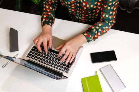 Female hand typing keyboard on laptop. Unrecognizable elegant woman working on computer at office. Start or finish of a work day. Workplace. Green notebook, phone. Top view.の写真素材