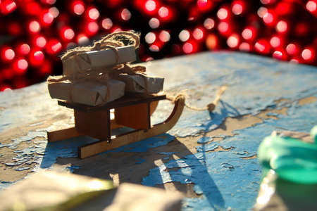 Christmas decoration. Wrapped craft vintage gift boxes lying wooden toy sled on blue old wooden background. Present box in craft paper. Holiday concept. Xmas backdrop. Many eco present. Red bokehの写真素材