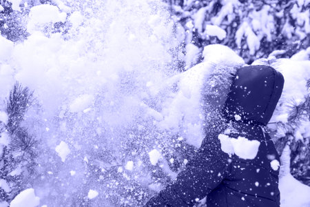 Preteen happy girl in pink warm hood shakes the snow from a branch outside on nature winter snowy forest background. Pretty child outdoor, cold weather. Color of the year 2022. Very peri tintの写真素材