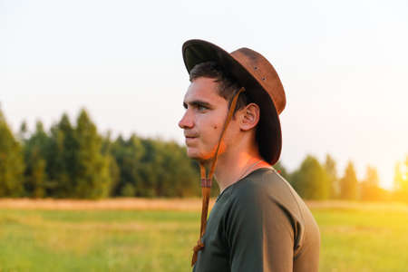 Young man farmer in cowboy hat at agricultural field on sunset with sun flare. Portrait of millennial man with hat, standing on nature background, outdoors in meadow. agriculture concept. copyspace.の写真素材