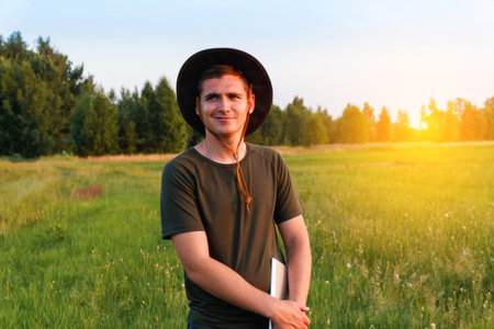 Young man farmer in cowboy hat at agricultural field on sunset holding tablet. Happy man on nature background, outdoors in meadow. Agriculture concept. Digital farmland. Smart worker. Overall.の写真素材
