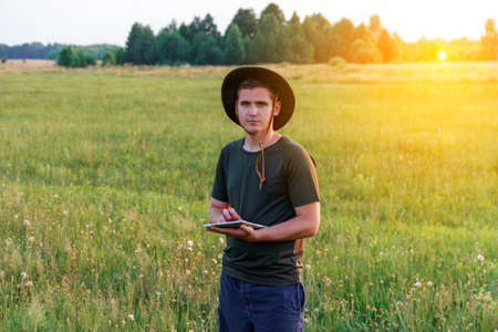 Young man farmer in cowboy hat at agricultural field on sunset holding tablet. Happy man on nature background, outdoors in meadow. Agriculture concept. Digital farmland. Smart worker. Overall.の写真素材