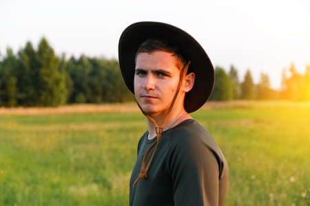 Young man farmer in cowboy hat at agricultural field on sunset with sun flare. Portrait of millennial man with hat, standing on nature background, outdoors in meadow. agriculture concept. copyspace.の写真素材