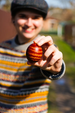 Happy smiling woman holding one colored red egg, Easter holiday concept. Hands holding modern painted easter eggs. Young hipster millennial woman in cap and retro clothes, british style. Pysanka.の写真素材