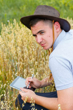 Young man smiling farmer in cowboy hat at agricultural field against blue sky. Portrait of...