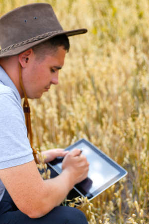 Digital farm. Portrait of farmer seating in gold wheat field and scrolling on tablet. Young man...