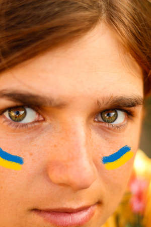 Ukrainian flag on the cheeks. Close-up portrait of a young woman 20, 25 years old. Green eyes, brown hair. Sunny, sunset, dawn. The concept of naturalness and tranquility. Selective focus.の写真素材