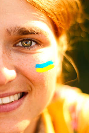 Ukrainian woman. Ukrainian flag on the cheeks. Selfie of a young woman 20, 25 years old. Half face, one green eye, brown hair. Sunny, sunset, dawn. Health and positive conceptの写真素材